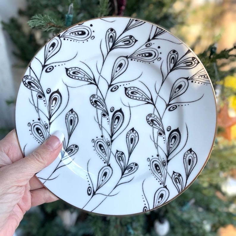 May include: White ceramic plate with a black and gold design. The plate features a repeating pattern of stylized black leaves and a gold rim. The design is hand-drawn and elegant, suitable for serving food or as a decorative piece.