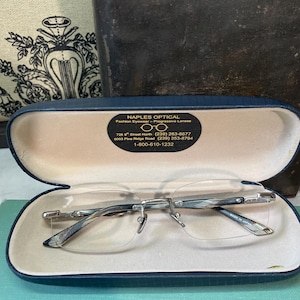 May include: Rimless eyeglasses with a silver frame rest inside a blue case with a beige interior. The case has the text "NAPLES OPTICAL" and contact information. The glasses are open, showcasing the lenses and frame details.