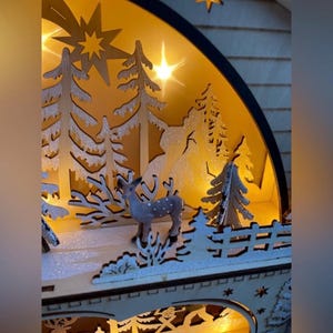 May include: A wooden Christmas scene with a deer, trees, and a bridge, illuminated by warm lights. The design features cut-out trees, a star, and a bridge, creating a festive holiday display.