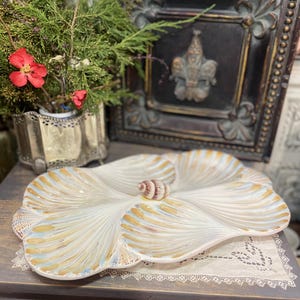 Puede incluir: Una fuente decorativa en forma de concha, de color blanco, con detalles dorados y azules. Una pequeña concha marina se encuentra en el centro. La fuente está sobre una superficie de madera con un tapete de encaje. Un elemento decorativo de pared enmarcado está en el fondo.