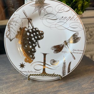 May include: White decorative plate with gold accents, featuring a grape cluster, dragonfly, and script that reads "Laughter". The plate is displayed on a gold-colored stand. The plate has the text "Receiving Joy from what is no" and "real Good seems but reas since we can".