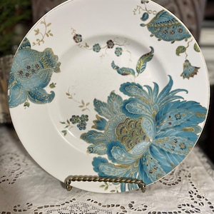 Puede incluir: Plato de cerámica blanca con un gran diseño floral turquesa detallado y ribete dorado. El plato presenta una flor central y pequeños detalles florales. El plato se exhibe sobre un soporte metálico decorativo.