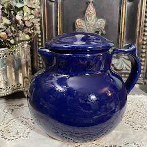 Puede incluir: Una jarra de cerámica azul cobalto con tapa. La jarra tiene un cuerpo redondeado, un asa y un pico. La tapa se asienta sobre la jarra. La jarra está sobre un mantel de encaje blanco.