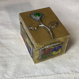 May include: A rectangular brass box with a rose design on the lid. The rose has a green gem center and silver-colored stem and leaves. The sides of the box feature colorful enamel panels with floral and figurative designs.