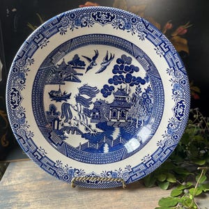 May include: A blue and white ceramic plate with a Willow pattern design. The plate features a detailed landscape scene with pagodas, bridges, and birds. The rim has a decorative border. The plate is displayed on a gold-colored stand.