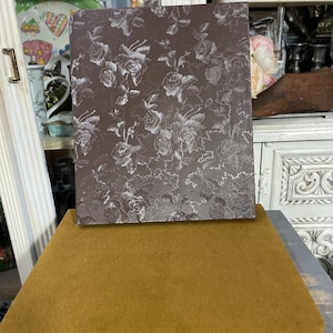 May include: A rectangular, brown floral-patterned book or decorative box rests on a mustard-colored, rectangular surface. The floral pattern features white roses. The base is a dark brown, rectangular shape.