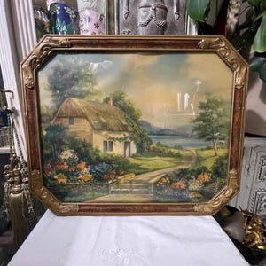 May include: An antique framed landscape painting featuring a thatched-roof cottage, a pathway, and a body of water. The painting has a warm color palette with a gold and brown ornate frame. The artwork depicts a serene countryside scene.