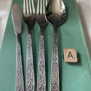 以下が含まれることがあります： バターナイフ、フォーク2本、スプーンを含む、花柄のヴィンテージカトラリーセット。銀色の食器は緑色の表面に置かれています。文字「A」と数字「1」が書かれた木製のスクラブルタイルも表示されています。