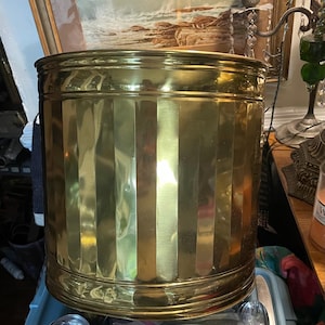 May include: A cylindrical brass wastebasket with vertical ribbed detailing. The wastebasket has a rounded top and bottom, and the brass has a polished finish. The wastebasket is a decorative item for a home or office.