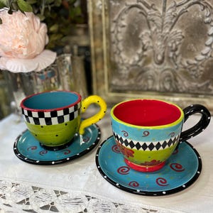 Könnte beinhalten: Zwei farbenfrohe Keramik-Teetassen und Untertassen. Eine Tasse hat einen grünen Körper mit schwarzen Punkten und einem karierten Rand, die andere hat ein Blumenmuster. Beide haben blaue Innenräume und stehen auf türkisfarbenen Untertassen mit schwarzen Rändern.
