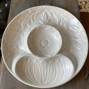 Puede incluir: Una fuente de cerámica blanca con tres secciones. La sección central es un cuenco pequeño, y la sección inferior es un cuenco más grande en forma de pétalo. El borde está decorado con un patrón similar a un helecho.