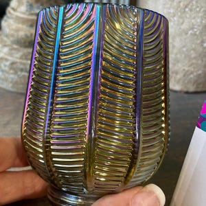 Puede incluir: Vaso de vidrio iridiscente con un diseño acanalado vertical texturizado. El vidrio refleja un espectro de colores, incluyendo oro, azul y púrpura. El vaso tiene una base redondeada y una parte superior ligeramente ensanchada, adecuado para bebidas.