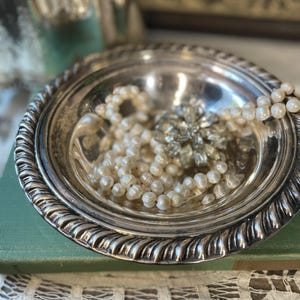 May include: A silver-colored, round dish with a decorative, twisted rope edge. The dish contains a pearl necklace and a silver-toned, floral-shaped brooch. The dish rests on a green book with a white lace trim.