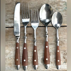 May include: A set of five stainless steel and wood-handled silverware pieces. The set includes a knife, two forks, and two spoons. The wooden handles are brown with a single white dot. The silverware is arranged on a wooden surface.