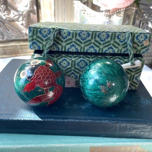 May include: Two green decorative spheres, one with a red fish design and the other with gold starburst accents, sit on a stack of books. A green and blue patterned box with handles is in the background. The spheres are likely made of stone.