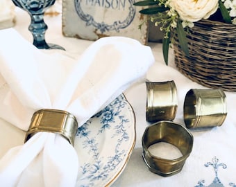 May include: A set of four brass napkin rings with a smooth, polished finish and a rounded rectangular shape. One ring secures a white cloth napkin on a blue and white floral patterned plate, while three additional rings sit nearby on the table.