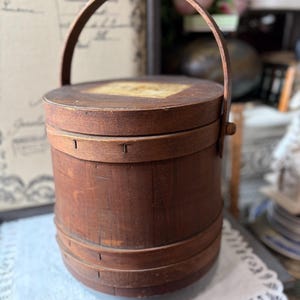 May include: A cylindrical wooden bucket with a dark brown finish, featuring a matching lid and a curved handle attached to the sides. The bucket is constructed with horizontal wooden bands and small metal fasteners visible along the upper rim.
