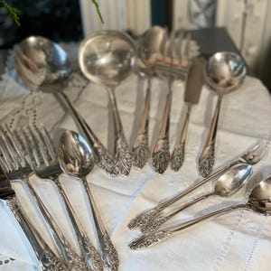 Puede incluir: Una colección de cubiertos antiguos, que incluye cucharas, tenedores y cucharones, dispuestos sobre un mantel de encaje blanco. Los cubiertos tienen detalles florales ornamentados en los mangos y una superficie brillante y reflectante. La disposición sugiere un entorno de comedor formal.