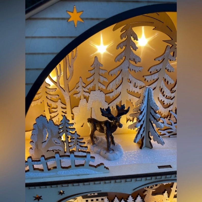 May include: A wooden Christmas scene with a moose figurine, snow-covered trees, and a star. The scene is illuminated by warm lights, creating a cozy, festive atmosphere. The design is intricate, with detailed cutouts.