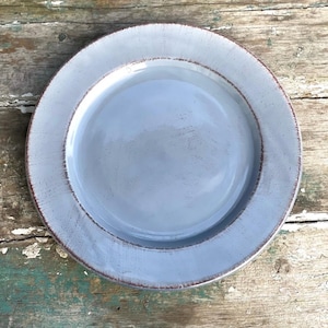 May include: A round, light blue ceramic plate with a slightly textured surface. The plate has a wide rim with a darker brown edge, creating a rustic, handcrafted appearance. The plate is designed for serving food.