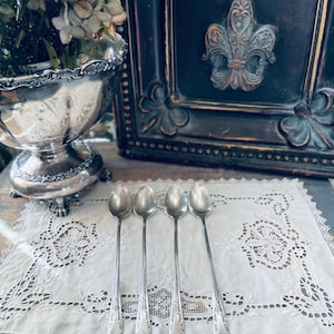 Può includere: Quattro cucchiai d'argento con manici lunghi poggiano su una tovaglietta di pizzo bianco. I cucchiai presentano dettagli decorativi sui manici. Una ciotola d'argento con fiori e un quadro scuro con un giglio sono sullo sfondo.