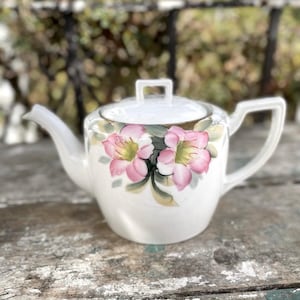 May include: White ceramic teapot with a lid and handle. The teapot is decorated with pink and green floral designs. The spout and handle are white. The teapot is sitting on a weathered wooden surface.
