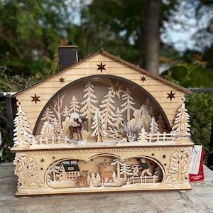 May include: A wooden Christmas-themed LED light-up house with a detailed forest scene. The house features cut-out trees, deer, and a snowy landscape. The house has a chimney and star cutouts, creating a warm, festive glow.