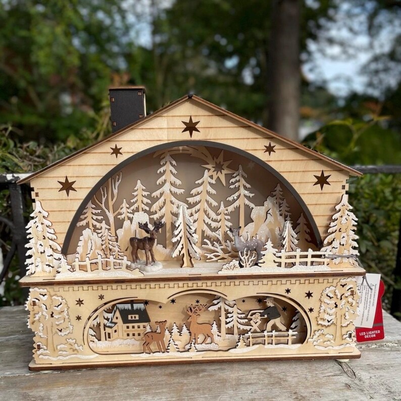 May include: A wooden Christmas-themed LED light-up house with a detailed forest scene. The house features cut-out trees, deer, and a snowy landscape. The house has a chimney and star cutouts, creating a warm, festive glow.