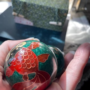 May include: Two green decorative spheres with a red and gold koi fish design. The spheres have a glossy finish and are held in a hand. A patterned box is in the background.