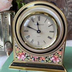 May include: A decorative, vintage "Wedgwood" quartz clock. The clock has a cream-colored face with black Roman numerals and hands. The clock is framed in gold and black, with a floral design at the base. The clock is made in England.