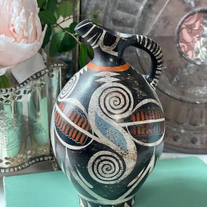 May include: A black ceramic pitcher with a handle and spout, adorned with white spiral and geometric designs, and orange and blue accents. The pitcher has a rounded body and a narrow neck. The design is reminiscent of ancient pottery.