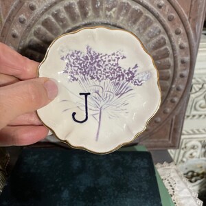 Puede incluir: Un peque&ntilde;o plato redondo de cer&aacute;mica blanquecina con borde dorado. El plato presenta un dise&ntilde;o floral morado y la letra "J" en azul oscuro. El plato est&aacute; sostenido en una mano.