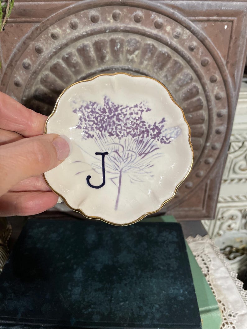 Puede incluir: Un peque&ntilde;o plato redondo de cer&aacute;mica blanquecina con borde dorado. El plato presenta un dise&ntilde;o floral morado y la letra "J" en azul oscuro. El plato est&aacute; sostenido en una mano.