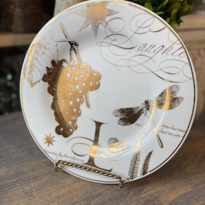 May include: A white decorative plate with gold detailing. The plate features a gold grape cluster, dragonfly, and star design. The word "Daughter" is written in gold script. The plate is displayed on a gold metal stand.