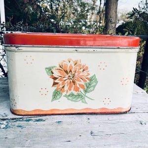 Op de afbeelding: Vintage rechthoekige metalen broodtrommel met een rood deksel en een crèmekleurige behuizing. De broodtrommel heeft een groot oranje en perzik bloemdesign met groene bladeren. Decoratieve oranje stippen en randen maken het ontwerp af.