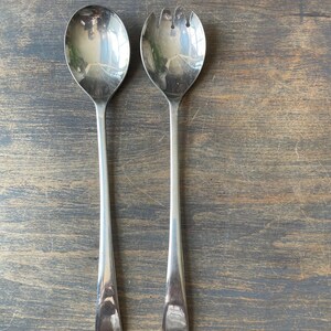 May include: A pair of silver serving utensils, comprising a large spoon and a fork with three tines. They have long, slender handles and are set on a dark, weathered wooden surface. The spoon and fork are reflective.