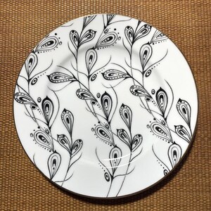 May include: White ceramic plate with a black and white floral design. The plate features a gold rim and a repeating pattern of stylized leaves and teardrop shapes. The design is intricate and detailed, creating a decorative and elegant aesthetic.
