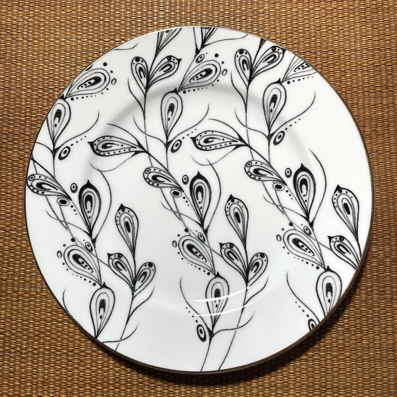 May include: White ceramic plate with a black and white floral design. The plate features a gold rim and a repeating pattern of stylized leaves and teardrop shapes. The design is intricate and detailed, creating a decorative and elegant aesthetic.
