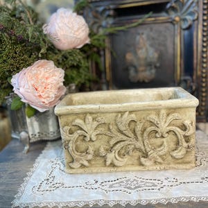 Peut inclure: Un pot rectangulaire beige clair avec des motifs floraux en relief. Le pot est posé sur un napperon en dentelle blanche. Des pivoines roses et un cadre décoratif sont en arrière-plan.