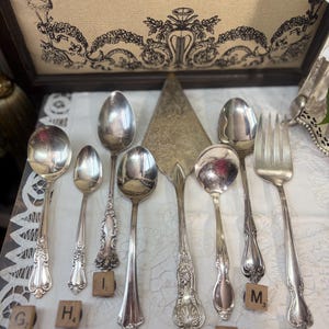 May include: A collection of eight silver-toned metal utensils including various spoons, a cake server, and a fork. Each piece features decorative embossed handles and is arranged with small wooden letter tiles labelled G, H, I, J, K, L, M, and N.
