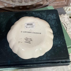 Puede incluir: Un peque&ntilde;o plato de baratijas Anthropologie de color blanquecino con un borde festoneado. El plato tiene una etiqueta con el nombre de la marca, un c&oacute;digo de producto y el precio. El texto "by ANTHROPOLOGIE" est&aacute; impreso en el plato.
