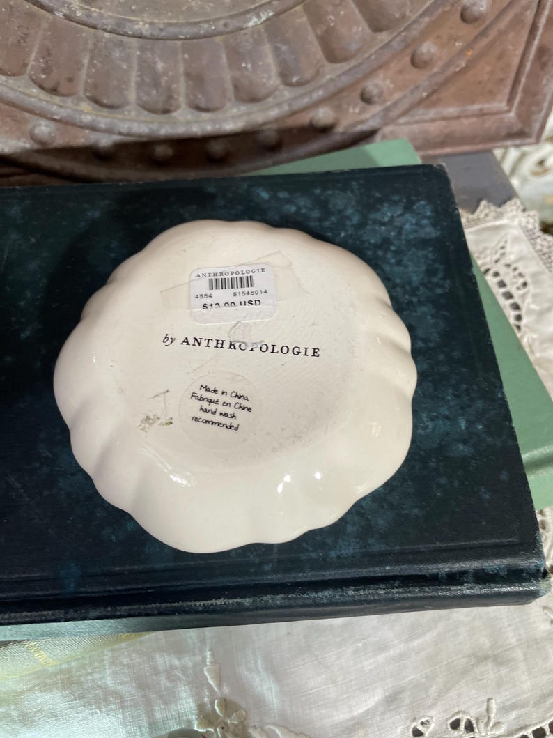 Puede incluir: Un peque&ntilde;o plato de baratijas Anthropologie de color blanquecino con un borde festoneado. El plato tiene una etiqueta con el nombre de la marca, un c&oacute;digo de producto y el precio. El texto "by ANTHROPOLOGIE" est&aacute; impreso en el plato.