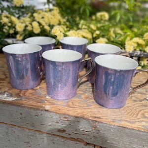 Peut inclure: Six tasses violettes irisées avec des anses dorées et des intérieurs blancs sont disposées sur une surface en bois vieilli. Les tasses ont une finition nacrée, reflétant des nuances de bleu et de rose. Des fleurs jaunes sont en arrière-plan.