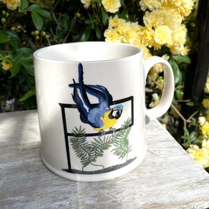 May include: White ceramic mug with a parrot illustration. The parrot has blue and yellow feathers and sits on a green leafy branch. The mug has a curved handle and is set against a backdrop of yellow flowers, typical of a garden.