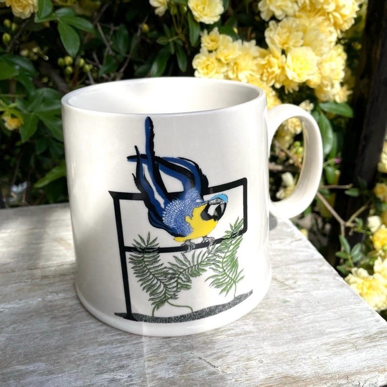 May include: White ceramic mug with a parrot illustration. The parrot has blue and yellow feathers and sits on a green leafy branch. The mug has a curved handle and is set against a backdrop of yellow flowers, typical of a garden.