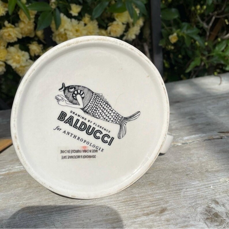 May include: A white ceramic mug featuring a black and white fish design and the text "BALDUCCI for ANTHROPOLOGIE". The mug is dishwasher and microwave safe.