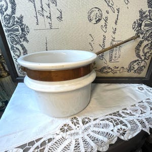 May include: A white ceramic pot with a wide, rounded rim and a smooth, glossy finish. A polished copper band encircles the upper section of the pot, and a long, slender metal handle extends from the side, providing a functional grip for the vessel.