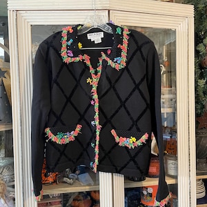 May include: Black cardigan sweater with a diamond pattern and colorful floral trim. The sweater has long sleeves, two pockets, and a collar with matching trim. The trim features pink, yellow, green, and blue accents. The sweater is hanging on a hanger.