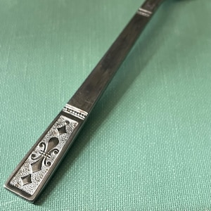 Può includere: Primo piano di un cucchiaio d'argento con manico decorativo. Il manico presenta un motivo a giglio in rilievo all'interno di una cornice rettangolare. Il cucchiaio poggia su una superficie strutturata verde chiaro.