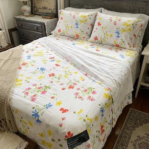 Pode incluir: Um conjunto de lençóis branco com um padrão floral colorido de flores amarelas, cor-de-rosa e azuis. O conjunto inclui duas fronhas e um lençol com uma etiqueta preta, indicando as dimensões de 205 por 244 cm.